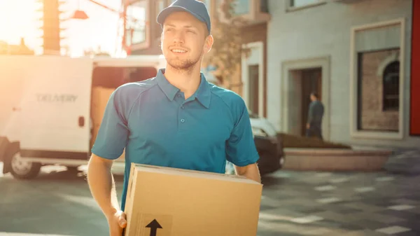 Portret van Delivery Man houdt Kartonnen doos Pakket Staande in Modern Stijlvol Business District met Bestelwagen in de achtergrond. Glimlachende koerier op weg naar postpakket leveren aan de klant. Zonnevlam — Stockfoto