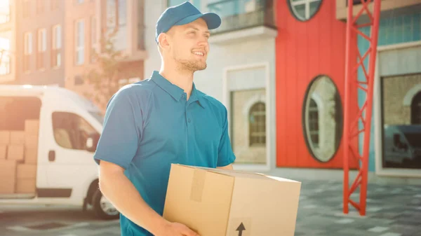 Portré of Delivery Man Holds kartondoboz csomag Álló modern üzleti negyed szállítási Van a háttérben. Mosolygó Futár Úton, hogy kézbesítse a postai csomagot az ügyfélnek. Napkitörés. — Stock Fotó
