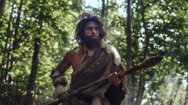 Tarih öncesi ormanda Hayvan Derisi Giyen İlkel Mağara Adamı 'nın Portresi ve Taş Ucu Mızrakla Kürk Avı. Tarih öncesi Neandertal Avcısı Ormanda İlkel Aletlerle Çöpleri Arıyor