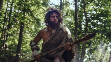 Portrait of Primeval Caveman Wearing Animal Skin and Fur Hunting with a Stone Tipped Spear in the Prehistoric Forest. Prehistoric Neanderthal Hunter Scavenging with Primitive Tools in the Jungle clipart