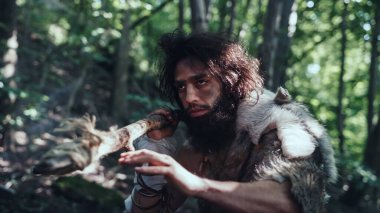 Tarih öncesi ormanda Hayvan Derisi Giyen İlkel Mağara Adamı 'nın Portresi ve Taş Ucu Mızrakla Kürk Avı. Tarih öncesi Neandertal Avcısı Ormanda İlkel Aletlerle Çöpleri Arıyor