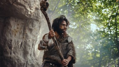 Hayvan Derisi Tutacağı Taşı Giyen İlkel Mağara Adamı Mağaradan çıkıyor ve Tarih Öncesi Araziye Bakıyor, Hayvan Avlamaya Hazır. Neanderthal Ormanda Avlanmaya Gidiyor