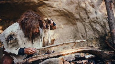 Hayvan derisi giyen ilkel mağara adamı yaylı matkapla ateş yakmaya çalışıyor. Neanderthal Kindle İnsan Uygarlığı tarihinde ilk insan yapımı yangın. Yemek pişirmek için ateş yakmak.