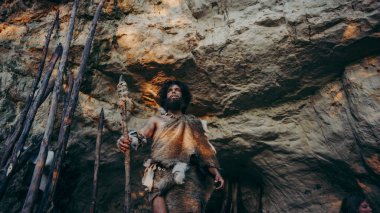 Hayvan Derisi ve Kürkü Taşı Tutan İlkel Mağara Adamı mağarasından avlanmaya hazır tarih öncesi ormana çıktı. Neanderthal Ormana Avlanmaya Gidiyor.