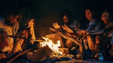 Neandertal ya da Homo Sapiens Ailesi Şenlik Ateşi Üzerinde Hayvan Eti Pişiriyor ve Sonra Onu Yiyor. Tarih öncesi avcı-toplayıcı kabilesi Gece vakti Karanlık Mağarada Hayvan Derileri Giyiyor