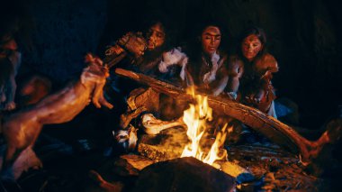 Neandertal ya da Homo Sapiens Ailesi Şenlik Ateşi Üzerinde Hayvan Eti Pişiriyor ve Sonra Onu Yiyor. Tarih öncesi avcı-toplayıcı kabilesi Gece vakti Karanlık Mağarada Hayvan Derileri Giyiyor