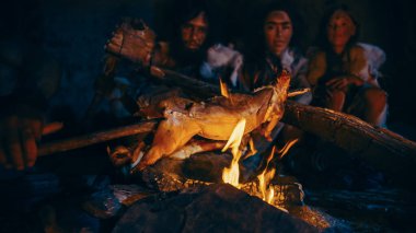 Neandertal ya da Homo Sapiens Ailesi Şenlik Ateşi Üzerinde Hayvan Eti Pişiriyor ve Sonra Onu Yiyor. Tarih öncesi avcı-toplayıcı kabilesi Gece vakti Karanlık Mağarada Hayvan Derileri Giyiyor.