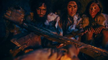 Neandertal ya da Homo Sapiens Ailesi Şenlik Ateşi Üzerinde Hayvan Eti Pişiriyor ve Sonra Onu Yiyor. Tarih öncesi avcı-toplayıcı kabilesi Gece vakti Karanlık Mağarada Hayvan Derileri Giyiyor.