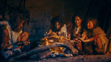 Neandertal ya da Homo Sapiens Ailesi Şenlik Ateşi Üzerinde Hayvan Eti Pişiriyor ve Sonra Onu Yiyor. Tarih Öncesi Avcı-Toplayıcılar kabilesi Hayvan Derisi Izgara Giyiyor ve Geceleri Mağarada Et Yiyiyor