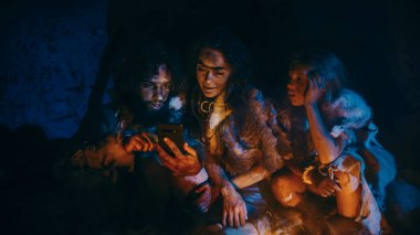 Tarih Öncesi, İlkel Avcı-Toplayıcılar kabilesi Hayvan Derisi Giyen Bir Mağarada Gece Akıllı Telefon Kullanıyor. Neandertal Homo Sapiens Ailesi Cep Telefonu 'nda İnternet Taraması, Ateşin Üzerinde Yemek Pişirme