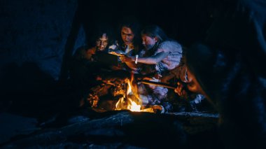 Tarih öncesi, İlkel Avcı-Toplayıcılar kabilesi Hayvan Derisi Giyen Geceleri Mağarada Dijital Tablet Bilgisayar Kullanıyor. Neanderthal ya da Homo Sapiens Ailesi İnternet Tarama, Video İzleme, TV Gösterileri