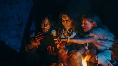Tarih öncesi, İlkel Avcı-Toplayıcılar kabilesi Hayvan Derisi Giyen Geceleri Mağarada Dijital Tablet Bilgisayar Kullanıyor. Neanderthal ya da Homo Sapiens Ailesi İnternet Tarama, Video İzleme, TV Gösterileri