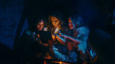 Tarih öncesi, İlkel Avcı-Toplayıcılar kabilesi Hayvan Derisi Giyen Geceleri Mağarada Dijital Tablet Bilgisayar Kullanıyor. Neanderthal ya da Homo Sapiens Ailesi İnternet Taraması, Video İzleme, Akım