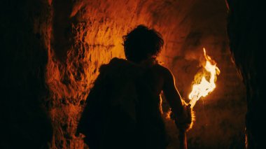 Primeval Caveman Wearing Animal Skin Exploring Cave At Night Holding Torch with Fire Looking at Drawings on the Walls at Night. Neanderthal Searching Safe Place to Spend the Night. Back View clipart