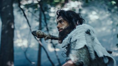 Hayvan Derisi Tutucu Taştan Mızraklı İlkel Mağara Adamı Etrafa Bakınıyor, Hayvan Avı Avında Tarih Öncesi Ormanı keşfediyor. Soğuk Işıkta Ormanda Avlanmaya Giden Neandertal