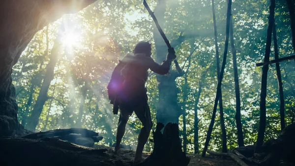 El cavernícola primitivo con piel de animal sostiene una lanza de punta de piedra, se encuentra en la entrada de la cueva mirando el bosque prehistórico listo para cazar presas de animales. Neanderthal va a cazar en la selva — Foto de Stock