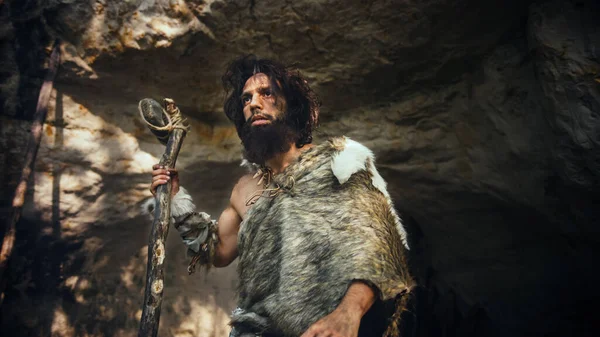 Primeval Caveman vestindo pele animal detém pedra derrubada martelo sai da caverna e olha em torno da floresta pré-histórica, pronto para caçar presas de animais. Neandertal vai caçar na selva . — Fotografia de Stock
