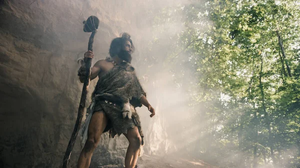 Primeval Caveman bär Animal Skin håller Stone Hammer står nära grottan och ser ut runt förhistoriska landskap, redo att jaga djur byte. Neandertalarnas jakt i djungeln. Lågvinkelskott — Stockfoto