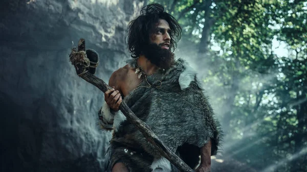 Primeval Caveman bär djur hud håller sten tippad hammare Kommer ut ur grottan och ser ut runt förhistoriska landskap, redo att jaga djur byte. Neandertalare på väg till jakten i djungeln — Stockfoto