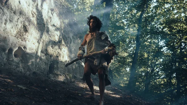 Primeval Caveman Wearing Animal Skin Holds Stone Tipped Hammer Comes out of the Cave and Looks Around, Exploring Prehistoric Forest Ready to Hunt Animal Prey. Neanderthal Going to Hunt in the Jungle