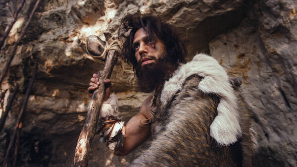 Primeval Caveman Wearing Animal Skin Holds Stone Hammer Looks Around Prehistoric Forest, Ready to Hunt Animal Prey. Neanderthal Going Hunting into the Jungle. Dramatic Low Angle Shot