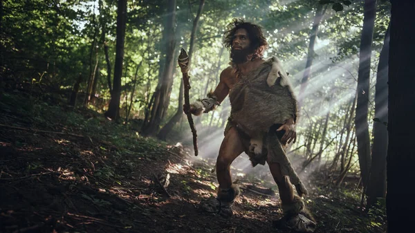 El cavernícola primitivo que lleva piel de animal sostiene la lanza de punta de piedra mira a su alrededor, explora el bosque prehistórico en una búsqueda de presas de animales. Neanderthal va a cazar en la selva — Foto de Stock