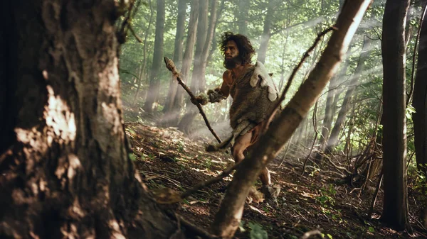 El cavernícola primitivo que lleva piel de animal sostiene la lanza de punta de piedra mira a su alrededor, explora el bosque prehistórico en una búsqueda de presas de animales. Neanderthal va a cazar en la selva — Foto de Stock