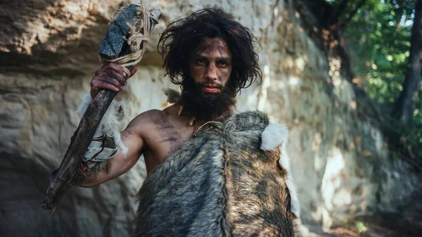Ősi ősember portréja, aki állati bőrt visel, kő hegyű kalapács. Őskori neandervölgyi vadász pózol primitív vadászattal a dzsungelben. Nézem a kamerát. — Stock Fotó