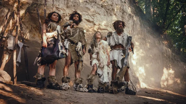 Négy vadász törzs, akik állati bőrt viselnek, kő hegyű szerszámokat hordanak, és a barlangjuk bejáratánál állnak. Két felnőtt férfi és két nőstény neandervölgyi portréja és életmódjuk — Stock Fotó