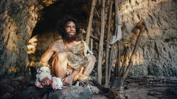 Pierwotny jaskiniowiec noszący zwierzęcą skórę trzyma kości i uderza w skałę. Neandertalczyk zabawiający się w pobliżu wejścia do jaskini, może tworząc pierwsze prymitywne narzędzia lub broń przez przypadek. — Zdjęcie stockowe