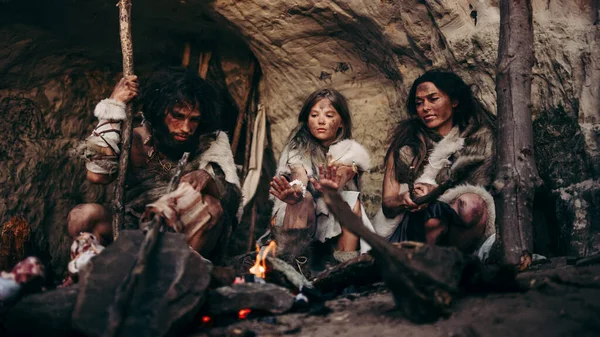 Племя доисторических первобытных охотников-собирателей, носивших анимированные шкуры, живет ночью в пещере. Семья неандертальцев или гомо сапиенсов пытается согреться у костра, держит руки над огнем, готовит еду — стоковое фото