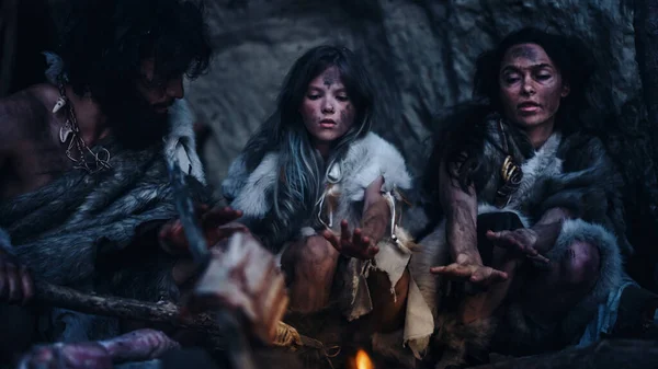 動物の皮を身に着けている先史時代のハンター・ギャザーの部族は夜に洞窟に住んでいます。ネアンデルタール人の家族はたき火で暖まり、火の上で手を握り、食べ物を調理しようとしている。暖かいフィルターで撮影. — ストック写真