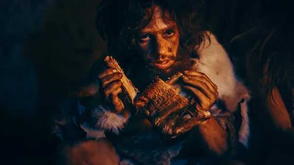 Primer plano Retrato del líder de la tribu usando piel de animal comiendo en una oscura cueva de miedo por la noche. Neandertal u Homo Sapiens Familia Cocinando Carne Animal sobre Hoguera y luego Comiéndolo . — Foto de Stock