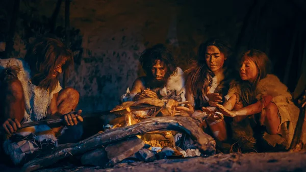 Neanderthal o Homo Sapiens Famiglia cucinare carne animale sopra il falò e poi mangiarlo. Tribù di cacciatori-raccoglitori preistorici che indossano pelli animali Grigliare e mangiare carne in grotta di notte — Foto Stock