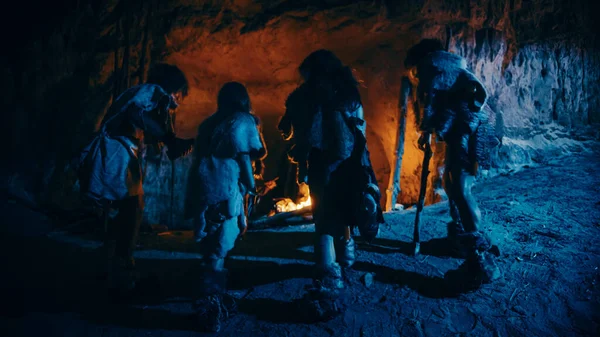 Tribe of Prehistoric Hunter-Gatherers Wearing Animal Skins Live in a Cave at Night (en inglés). Neanderthal o la familia Homo Sapiens tratando de calentarse en la hoguera, sosteniendo las manos sobre el fuego. Vista trasera — Foto de Stock