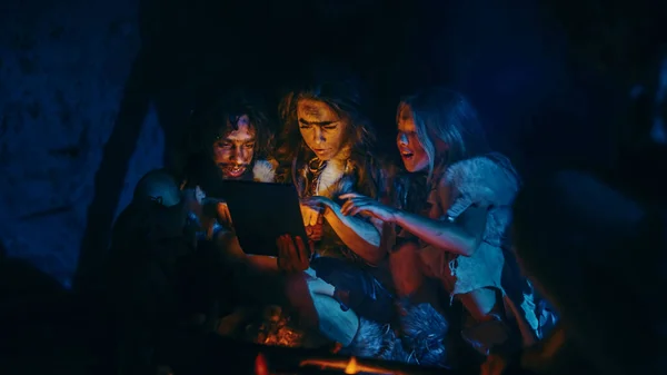 Tribe of Prehistoric, Primitive Hunter-Gatherers Wearing Animal Skins Use Digital Tablet Computer in a Cave at Night. Neanderthal or Homo Sapiens Family Browsing Internet, Watching Videos, Streaming