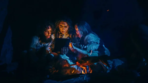 Der Stamm der prähistorischen, primitiven Jäger und Sammler, die Tierhäute tragen, nutzt nachts einen digitalen Tablet-Computer in einer Höhle. Neandertaler oder Homo-Sapiens-Familie surft im Internet, schaut Videos, streamt — Stockfoto