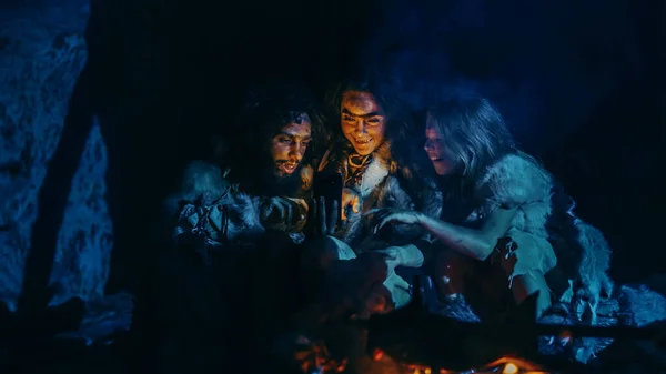 Tribe of Prehistoric, Primitive Hunter-Gatherers Wearing Animal Skins Use Smartphone in a Cave at Night (en inglés). Neanderthal Homo Sapiens Familia Navegación por Internet en el teléfono móvil — Foto de Stock