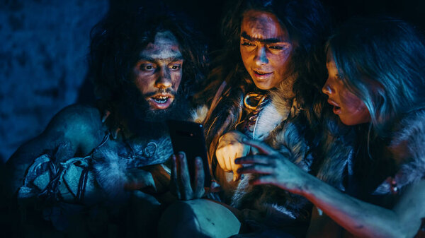 Tribe of Prehistoric, Primitive Hunter-Gatherers Wearing Animal Skins Use Smartphone in a Cave at Night. Neanderthal Homo Sapiens Family Browsing Internet on Mobile Phone