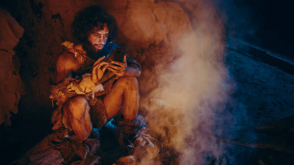 Tribe of Prehistoric, Primitive Hunter Gatherer Wearing Animal Skin Uses Smartphone in a Cave at Night. Neanderthal Homo Sapiens Male Browsing Internet on Mobile Phone, Watches Videos