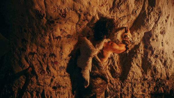 Primitive Prehistoric Neanderthal Wearing Animal Skin Draws Animals and Abstracts on the Walls at Night. Creating First Cave Art with Petroglyphs, Rock Paintings Illuminated by Fire. Back View