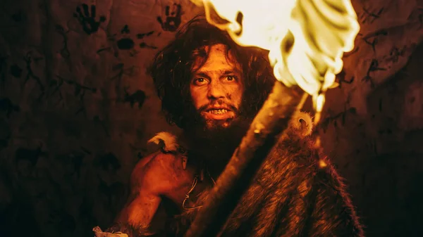 Retrato del hombre de las cavernas primitivo con piel de animal de pie en su cueva por la noche, sosteniendo la antorcha con fuego. Primitivo Neanderthal Hunter Homo Sapiens por la noche solo. En el fondo Dibujos de arte de la cueva — Foto de Stock