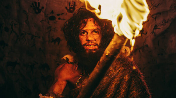 Portrait of Primeval Caveman Wearing Animal Skin Standing in His Cave At Night, Holding Torch with Fire. Primitive Neanderthal Hunter Homo Sapiens At Night Alone. In the Background Cave Art Drawings