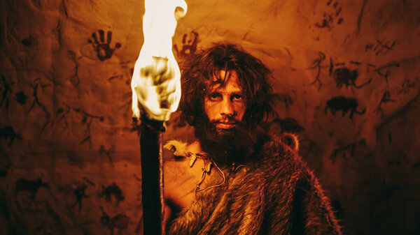 Portrait of Primeval Caveman Wearing Animal Skin Standing in His Cave At Night, Holding Torch with Fire. Primitive Neanderthal Hunter Homo Sapiens At Night Alone. In the Background Cave Art Drawings
