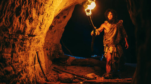 Primeval Caveman Wearing Animal Skin Exploring Cave At Night, Holding Torch with Fire Looking at Drawings on the Walls at Night. Neanderthal Searching Safe Place to Spend the Night