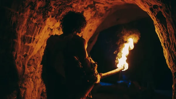 Pierwotny jaskiniowiec noszący skórę zwierzęcia stoi w jaskini w nocy, trzymając pochodnię z ogniem wyglądającym nocą przez jaskinię. Powrót Widok — Zdjęcie stockowe