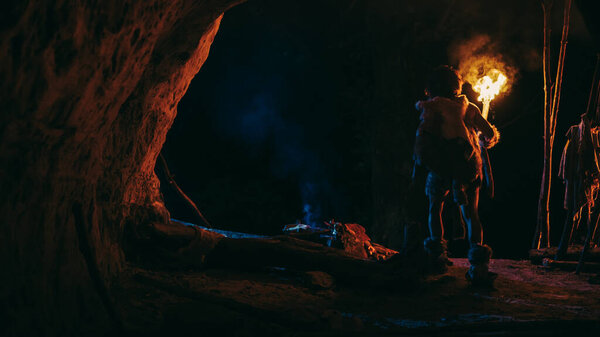 Primeval Caveman Wearing Animal Skin Stands in a Cave At Night, Holding Torch with Fire Looking Out of The Cave at Night. Back View