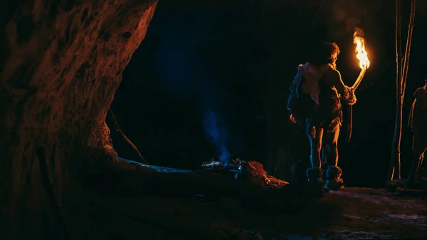 Hombre de las cavernas primitivo con piel de animal se para en una cueva por la noche, sosteniendo la antorcha con fuego mirando hacia fuera de la cueva por la noche. Vista trasera — Foto de Stock