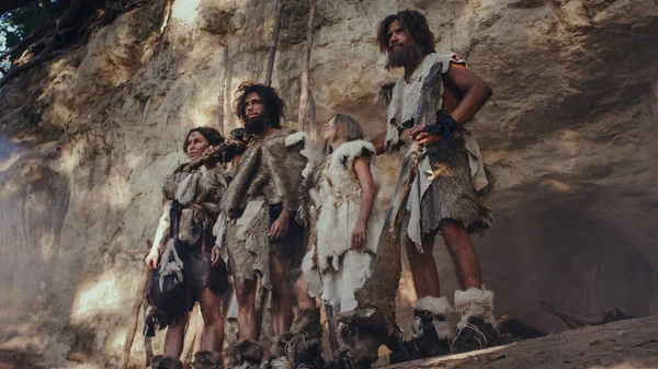 Племя охотников-собирателей, носящее шкуру животного, держащее каменные наконечники, стоит у входа в пещеру. Семья неандертальцев готова к охоте в джунглях или миграции — стоковое фото