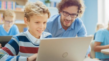 İlköğretim Okulu Bilgisayar Bilimi Sınıfı: Akıllı Çocuk Laptop Bilgisayar Kullanıyor, Arkadaş Canlısı Öğretmen Ders Açıklayarak Ona Yardım Ediyor. Çocuklar BTMM 'de Modern Eğitim Alırken, Oynarken ve Öğrenirken